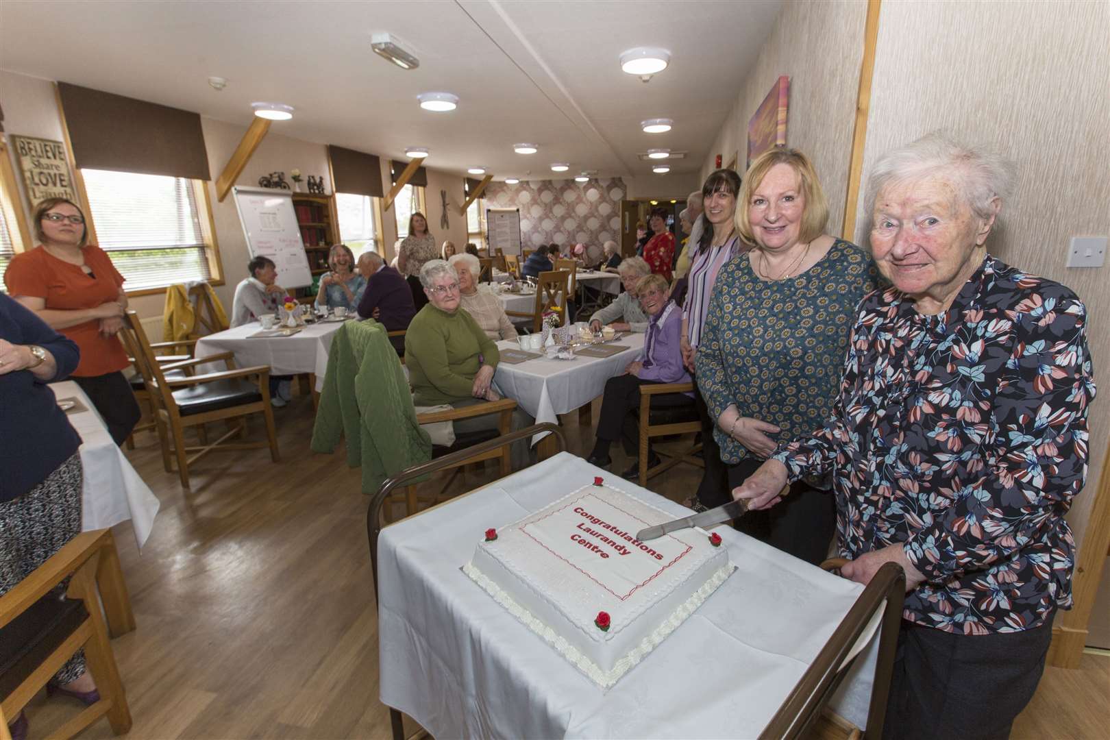 Dorrie Barnes (98), the centre's oldest service user, cuts a cake to mark the occasion, watched by centre manager Margaret Allan and some of the guests, staff and attendees. Picture: Robert MacDonald / Northern Studios