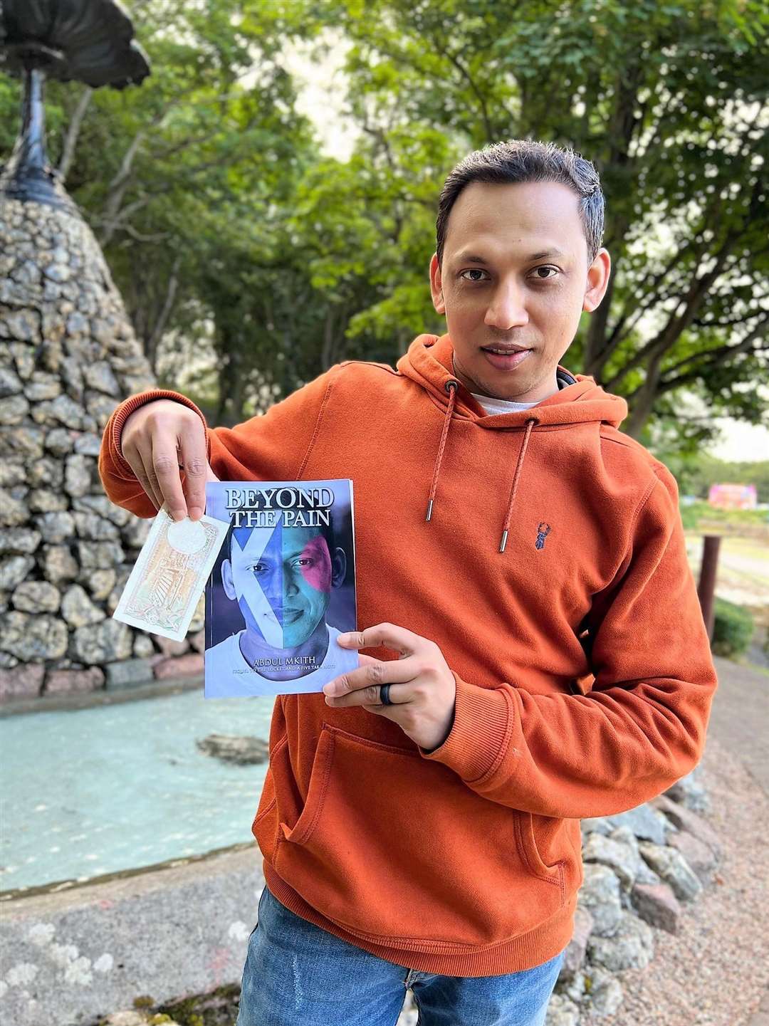 Abdul Mkith with his new book Beyond the Pain and holding a Bangladeshi five taka note. He will give out the notes for readers to use as bookmarks.