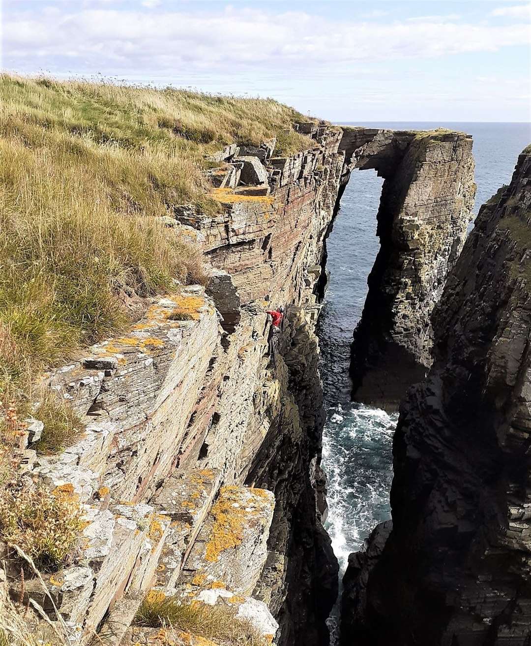 A climber in red can be seen scaling the cliff face near the Brig of Stack.