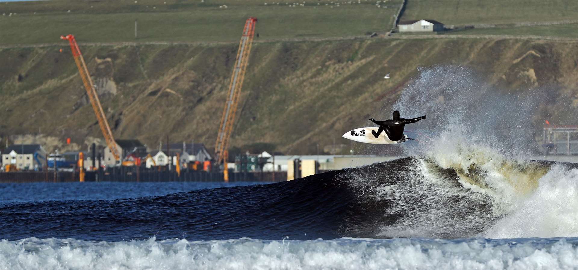 A surfer takes to the air at Thurso East, with cranes working on the new pier at Scrabster in the background. Picture: James Gunn