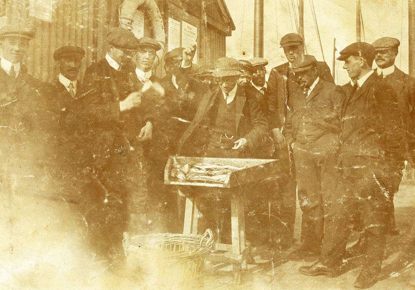 We think this photo of a herring auction on the Wick quayside was taken in about 1910. A sample of fish from one boat’s catch is on the table and the auctioneer, hand raised, is calling for bids for the whole catch from the onlooking fishcurers. (Andy Anderson)